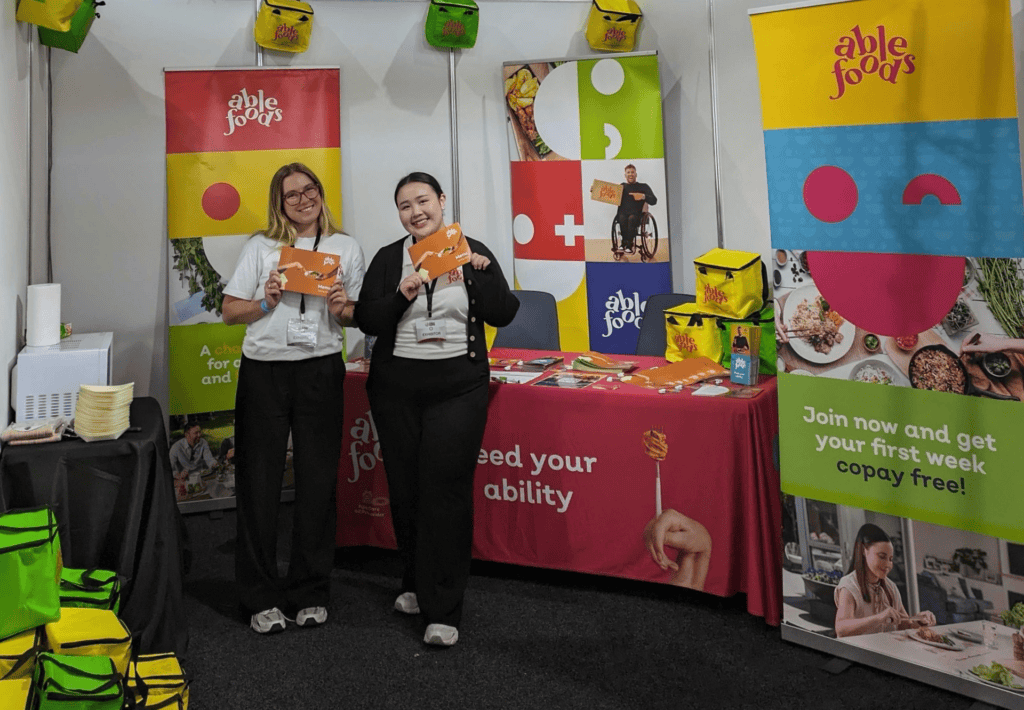 A blonde woman and an Asian woman holding orange Able Foods menus, in front of our display of freebies and colourful pull-up banners.