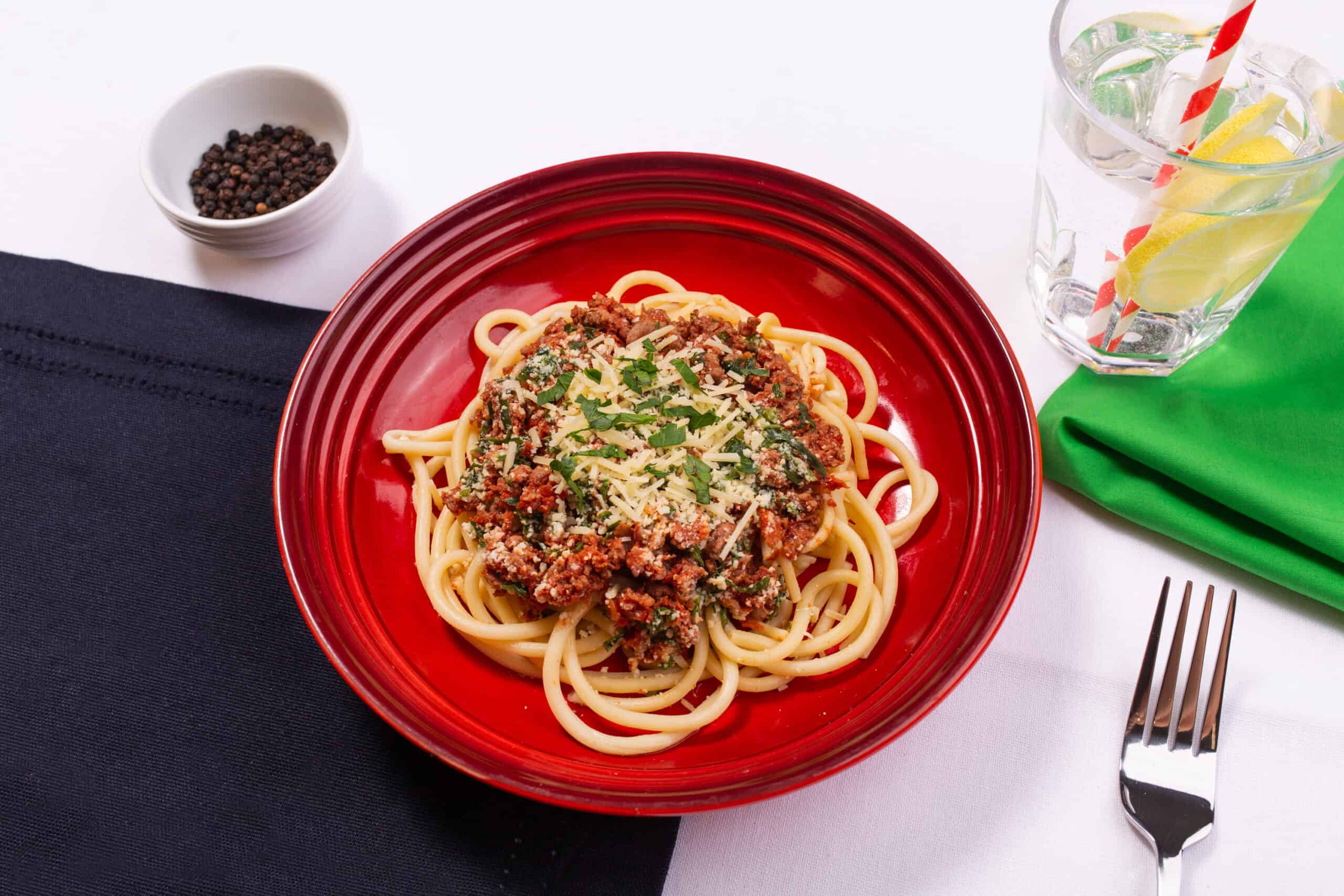 spaghetti bolognese on a red plate, with a water glass, green napkin, and fork in the background