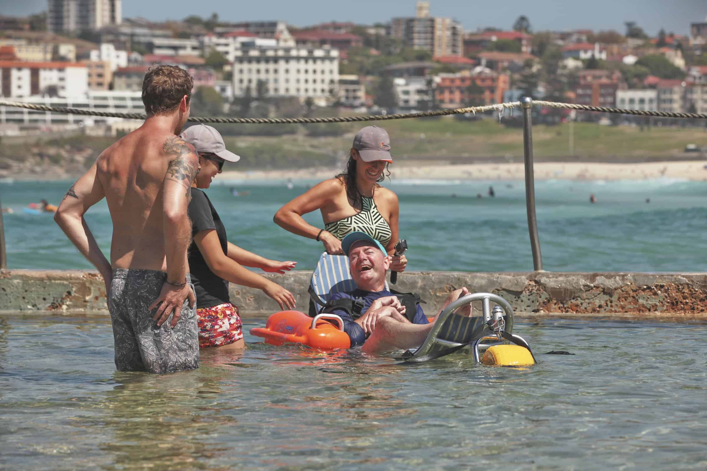 man in mobility beach chair sitting in the ocean, with three people standing around him